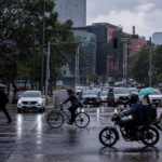 Automóviles avanzan lentamente sobre una avenida de la Ciudad de México mientras peatones cruzan con paraguas bajo un cielo nublado; al fondo se observan señales viales y edificios urbanos.