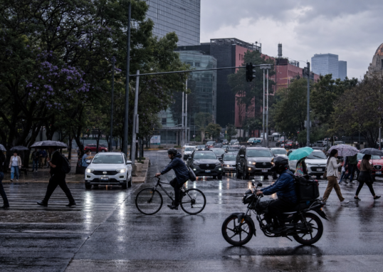 Automóviles avanzan lentamente sobre una avenida de la Ciudad de México mientras peatones cruzan con paraguas bajo un cielo nublado; al fondo se observan señales viales y edificios urbanos.