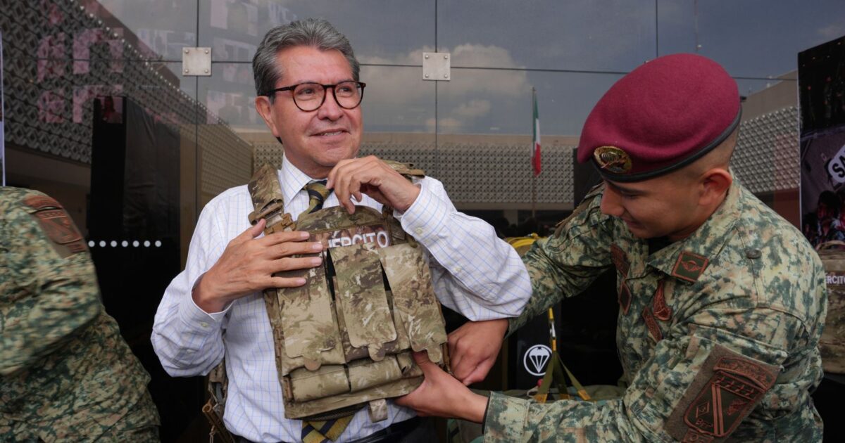 Ricardo Monreal ajusta un chaleco táctico con ayuda de un militar durante una exhibición.