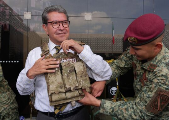Ricardo Monreal ajusta un chaleco táctico con ayuda de un militar durante una exhibición.