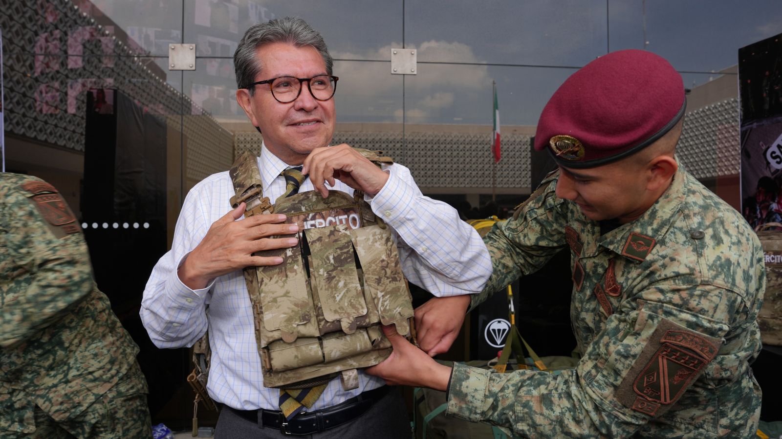 Ricardo Monreal ajusta un chaleco táctico con ayuda de un militar durante una exhibición.