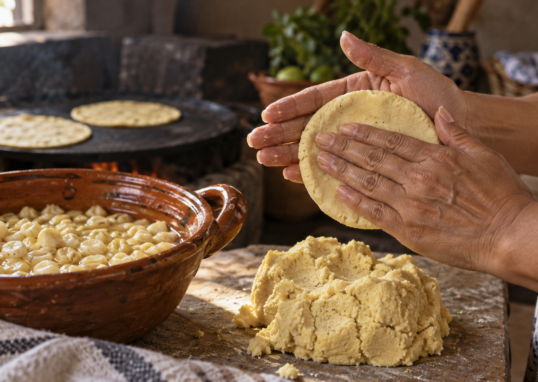 Manos formando una tortilla de masa junto a maíz cocido, con un comal al fondo; ilustración generada con IA.