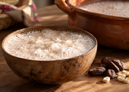 Jícara con bebida beige y espuma blanca grumosa, con granos de cacao y maíz sobre una mesa; ilustración generada con IA.
