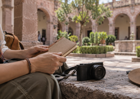 Manos con libreta y cámara sin marcas ante un patio de arquería desenfocado; ilustración conceptual generada con IA.